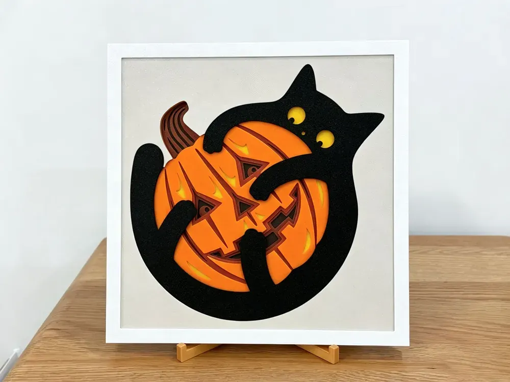 Tableau d’Halloween en couches : un chat noir aux yeux jaunes enlaçant une citrouille sculptée orange sur un panneau crème, dans un cadre blanc, sur un support