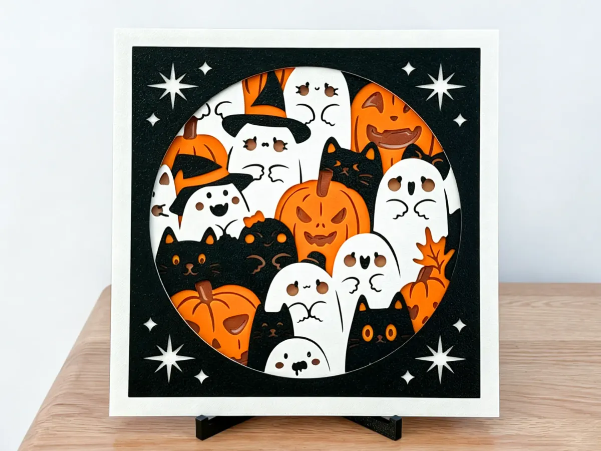 Tableau d’Halloween en couches : citrouilles, fantômes et chats noirs dans une fenêtre ronde, bordure blanche et panneau noir aux étoiles découpées, sur un support