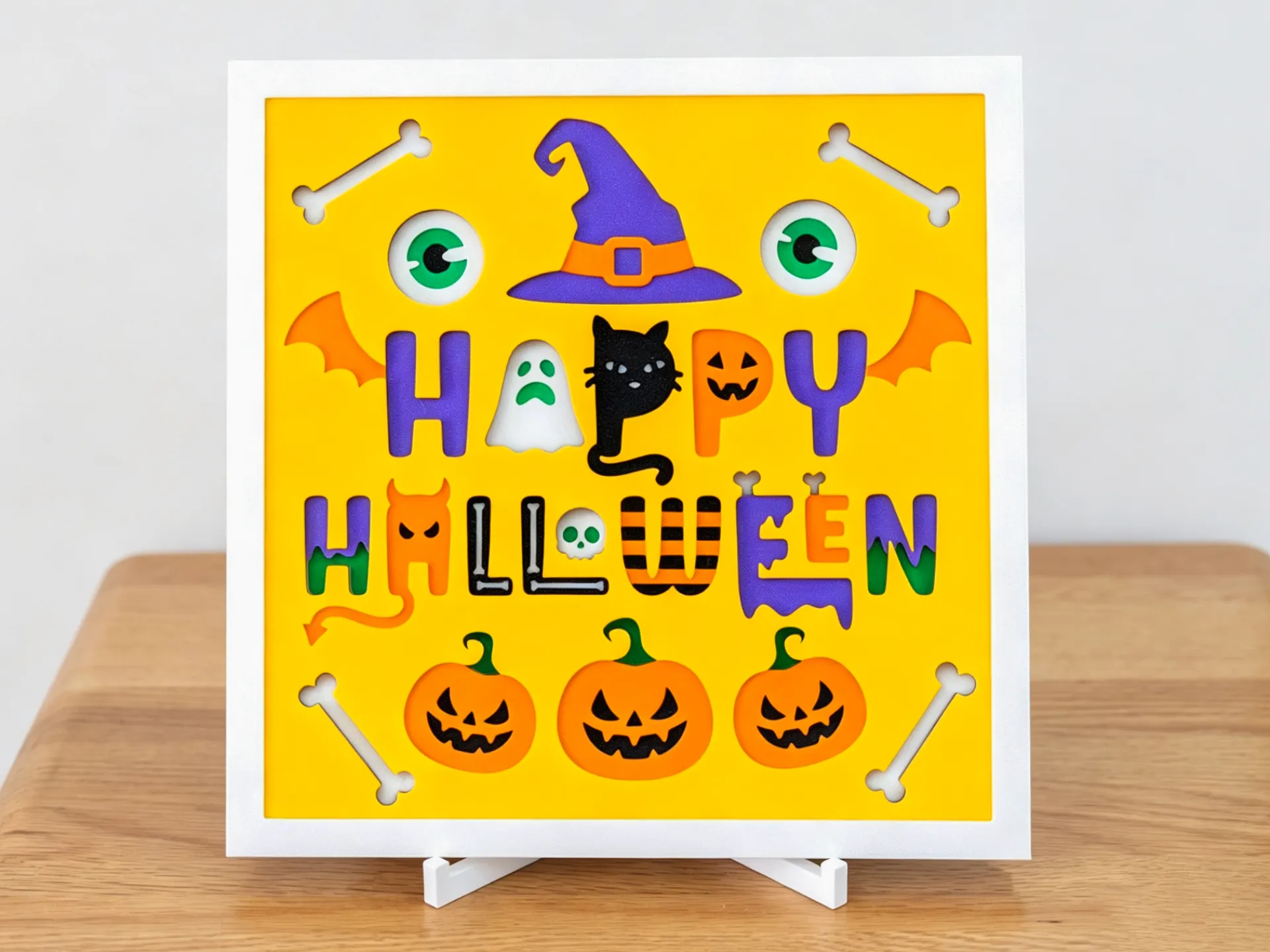 Happy Halloween layered art: colourful lettering with a ghost, a black cat, pumpkins, a witch hat and bones on a yellow panel in a white frame, on a stand